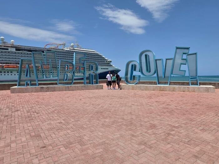 Travelers posing by the Amber Cove sign with a cruise ship behind them